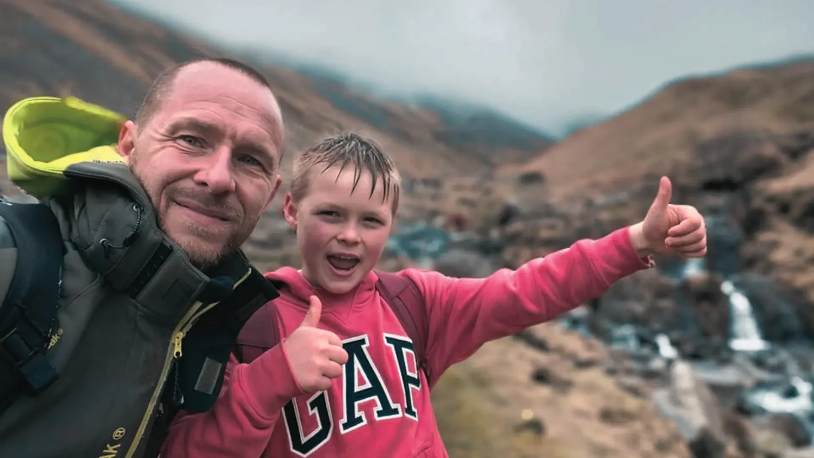 Micheal Hilton smiling on a hill walk beside a waterfall, next to a young boy giving a thumbs up